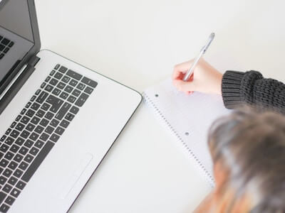 Website launch checklist for WordPress. Top down image of a fair skinned person writing on a ruled notebook with a pen. Next to the notebook is a silver coloured laptop with black keyboard.