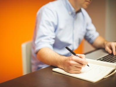Website launch checklist for WordPress. Image of a fair-skinned person in a shirt writing on a notebook with their right hand, and typing on a silver coloured laptop with their left hand. They are seated on a white chair, and using a dark wooden desk. The background is bright orange.